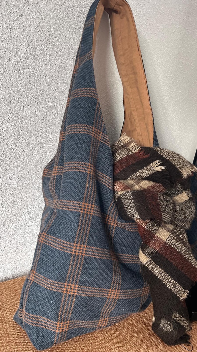 May include: A hobo-style shoulder bag made of blue fabric with an orange plaid pattern, featuring a wide strap and a tan interior lining. A folded scarf with a brown, cream, and dark red plaid pattern is tucked inside the bag's main opening.