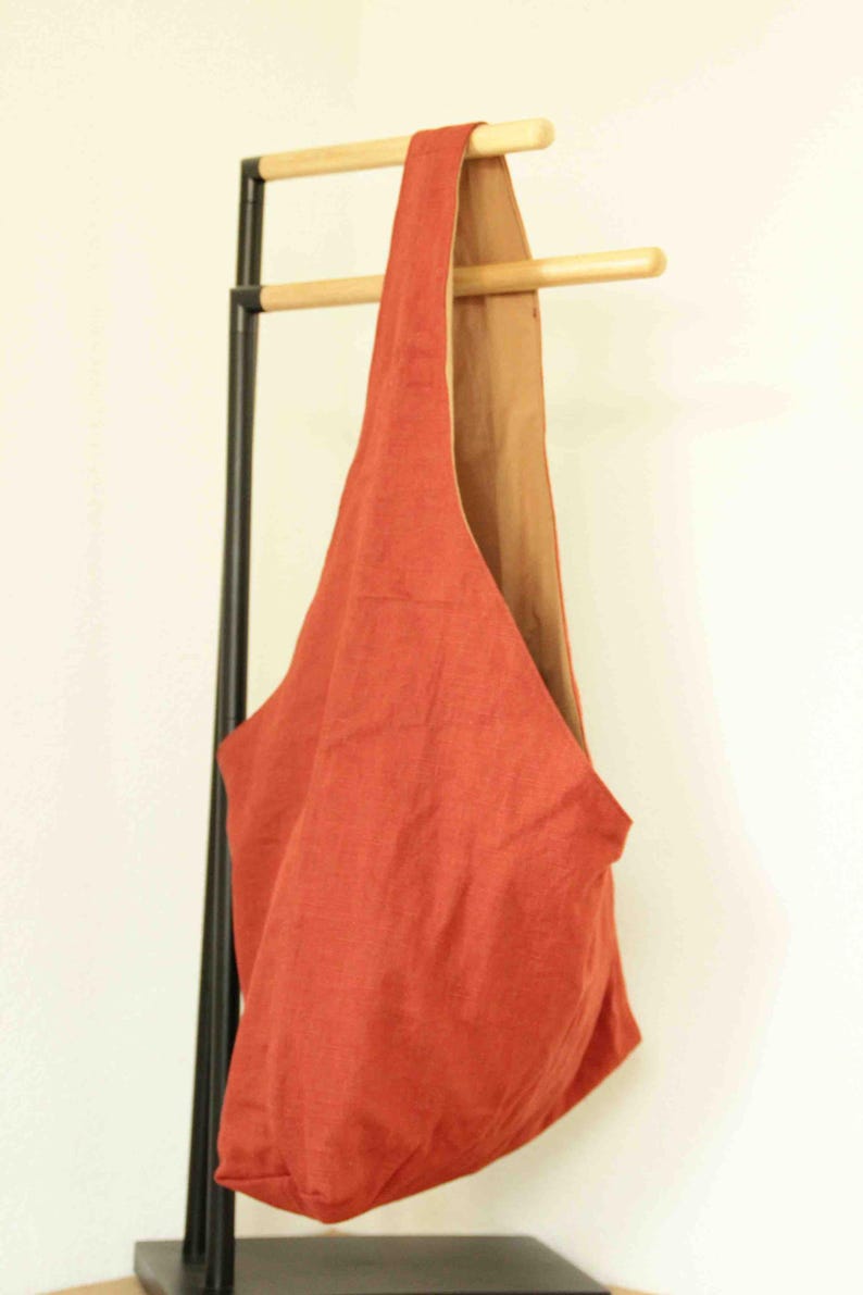 May include: A hobo-style shoulder bag featuring a vibrant rust-orange exterior and a contrasting tan interior lining. The bag has a single integrated strap design that forms a continuous loop, creating a soft, unstructured silhouette for carrying.