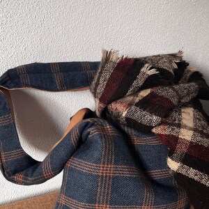 May include: A blue and orange plaid hobo bag with a wide shoulder strap and a tan leather-like interior lining, paired with a folded plaid scarf featuring a dark brown, cream, and deep red checkered pattern with frayed edges.
