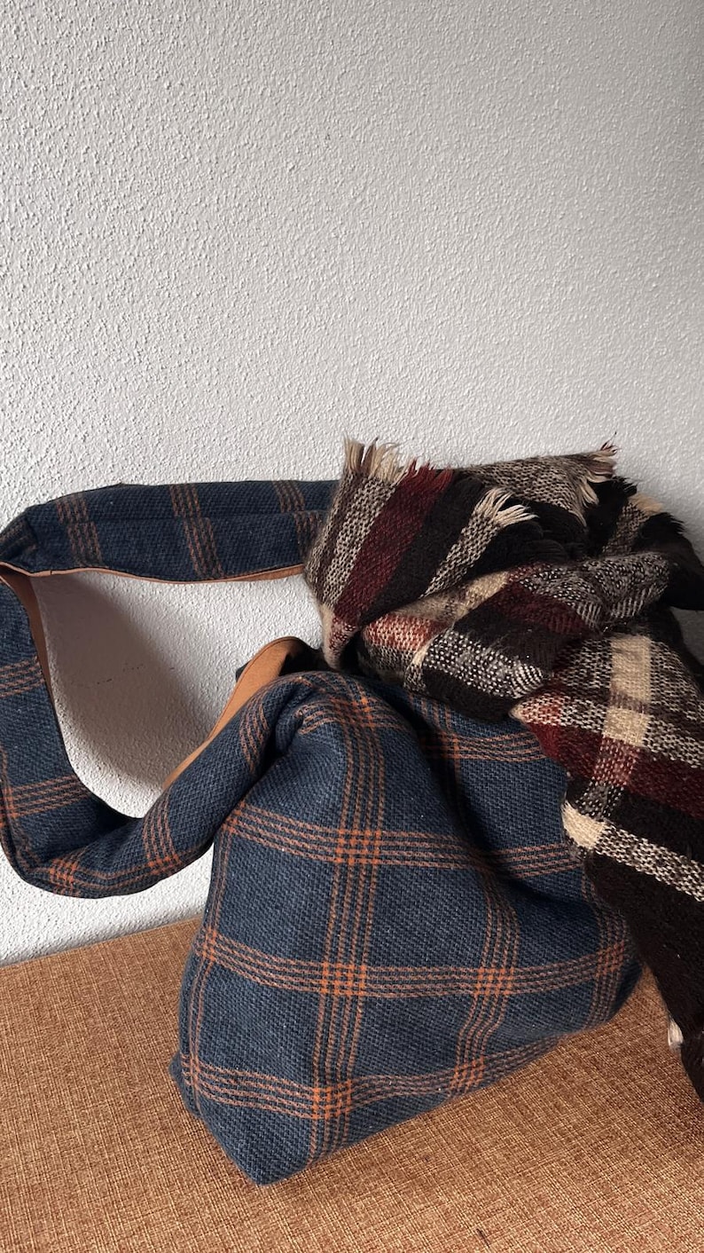 May include: A blue and orange plaid hobo bag with a wide shoulder strap and a tan leather-like interior lining, paired with a folded plaid scarf featuring a dark brown, cream, and deep red checkered pattern with frayed edges.