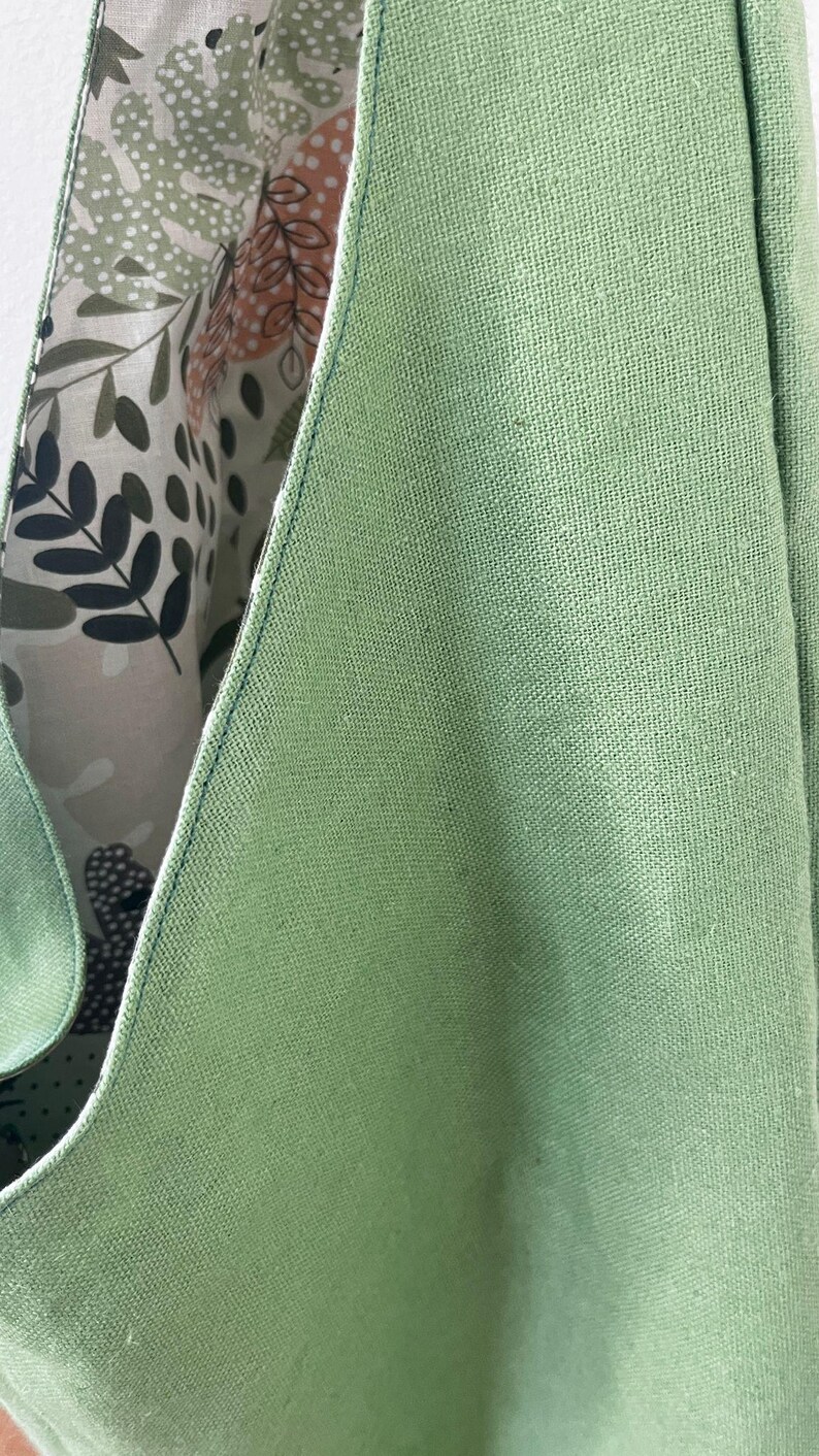 May include: A sage green tote bag with a textured fabric exterior and a contrasting interior lining featuring a botanical print of leaves, stems, and abstract shapes in shades of dark green, light green, and muted orange on a cream background.