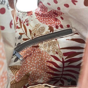 May include: The interior of a fabric bag featuring a cream-colored lining with a repeating pattern of stylized leaves, dots, and botanical shapes in shades of terracotta, brown, and gray. It includes a horizontal zippered pocket with a gray zipper.