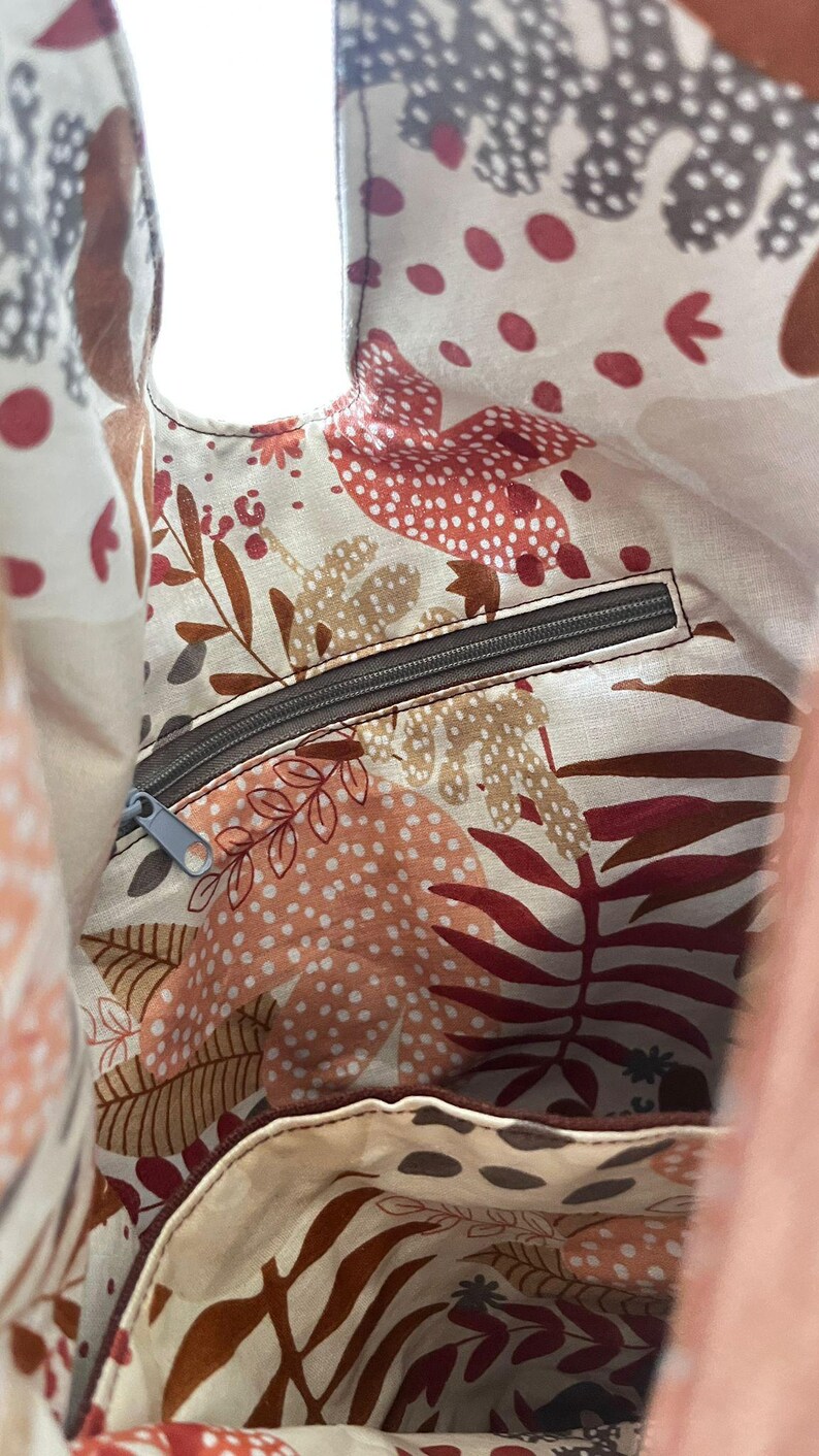 May include: The interior of a fabric bag featuring a cream-colored lining with a repeating pattern of stylized leaves, dots, and botanical shapes in shades of terracotta, brown, and gray. It includes a horizontal zippered pocket with a gray zipper.