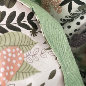 May include: A fabric item featuring a light pink background with a botanical print of dark green leaves, black fern silhouettes, and peach-colored dotted circles. The edge is finished with a solid sage green fabric trim and visible stitching.