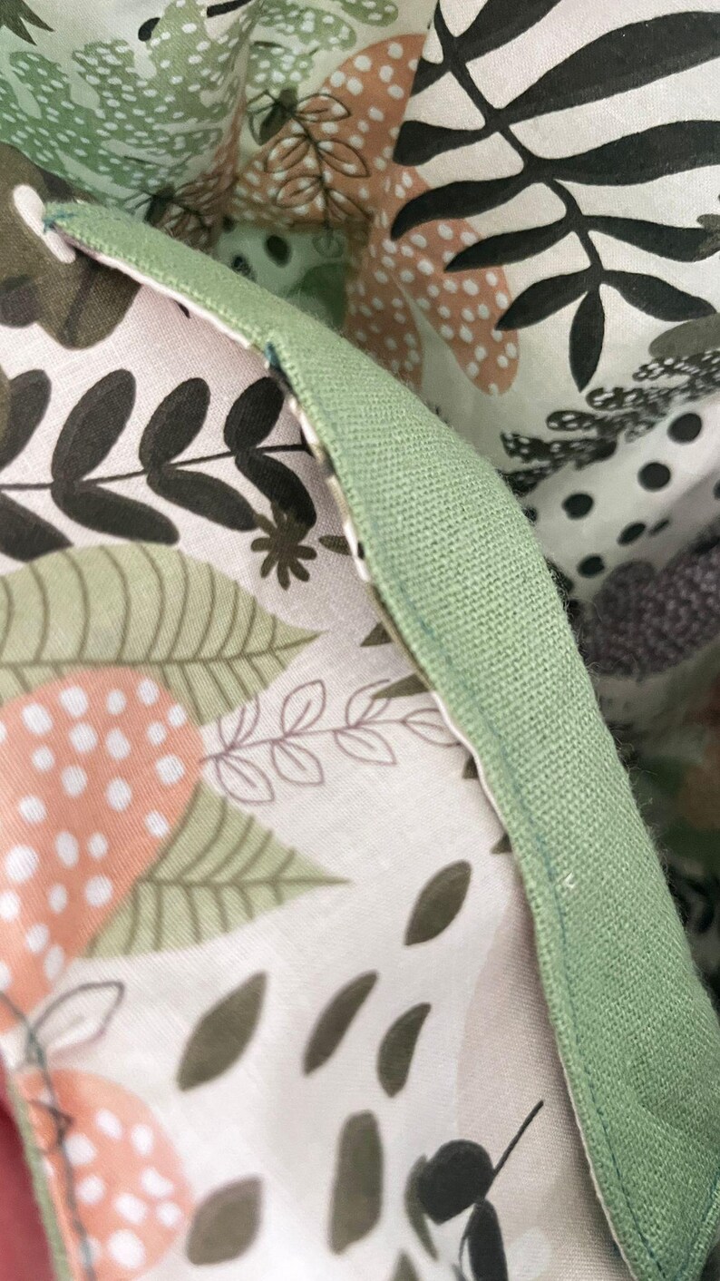 May include: A fabric item featuring a light pink background with a botanical print of dark green leaves, black fern silhouettes, and peach-colored dotted circles. The edge is finished with a solid sage green fabric trim and visible stitching.