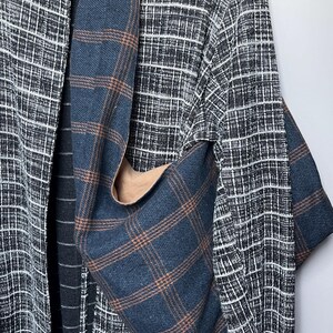 May include: A hobo bag made of blue fabric with a brown plaid pattern hanging over a black and white textured open-front cardigan. The bag features a wide shoulder strap and a slouchy, unstructured shape, while the cardigan has a grid pattern.