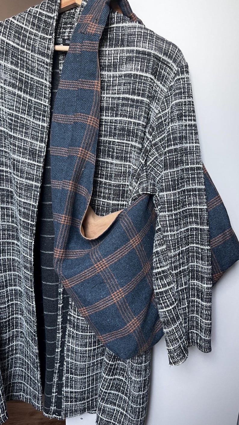 May include: A hobo bag made of blue fabric with a brown plaid pattern hanging over a black and white textured open-front cardigan. The bag features a wide shoulder strap and a slouchy, unstructured shape, while the cardigan has a grid pattern.