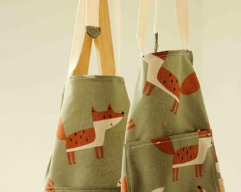 May include: Two sage green aprons featuring a repeating pattern of stylized orange and white foxes with black spots. Each apron includes light beige fabric neck straps and a front pocket on the right side, displaying the fox print design.
