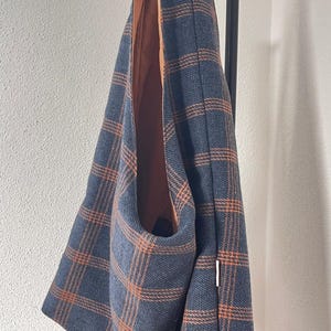 May include: A hobo-style shoulder bag featuring a dark blue and orange plaid pattern. The bag has a single integrated strap and a solid orange interior lining. A small white rectangular fabric tag is visible on the side seam of the exterior.