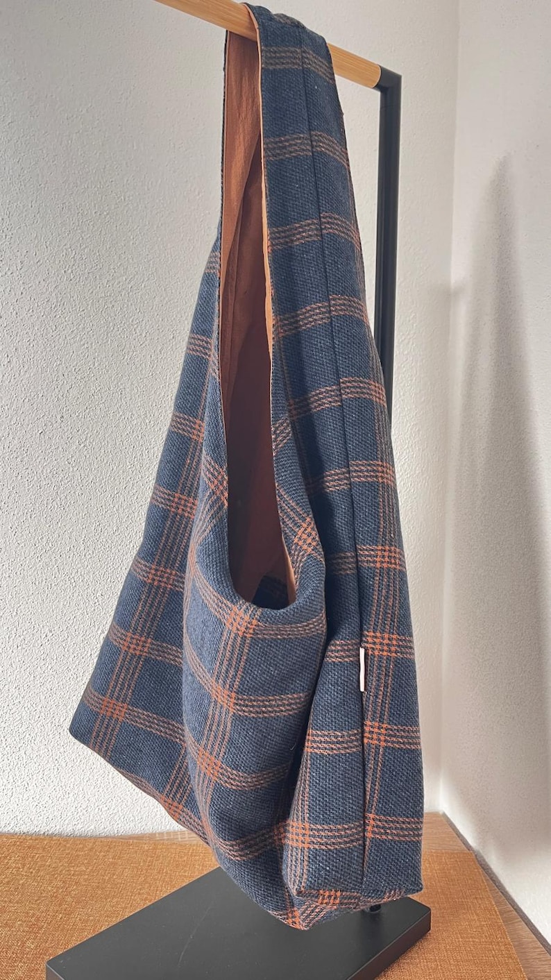 May include: A hobo-style shoulder bag featuring a dark blue and orange plaid pattern. The bag has a single integrated strap and a solid orange interior lining. A small white rectangular fabric tag is visible on the side seam of the exterior.