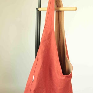 May include: A solid burnt orange hobo bag with a single wide shoulder strap and a contrasting tan interior lining. The bag features a simple, unstructured design with a small white rectangular tag attached to the side seam, hanging from a wooden rack.