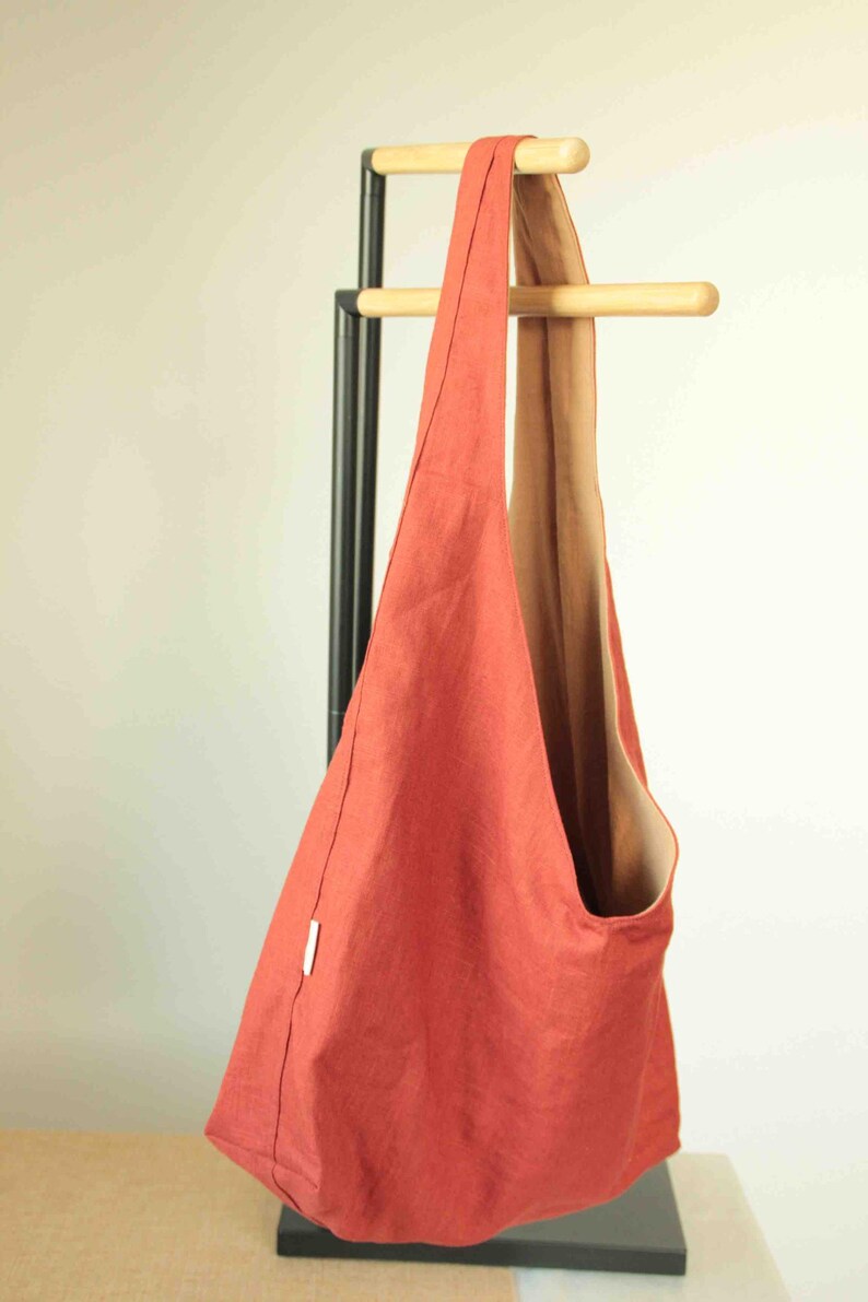 May include: A solid burnt orange hobo bag with a single wide shoulder strap and a contrasting tan interior lining. The bag features a simple, unstructured design with a small white rectangular tag attached to the side seam, hanging from a wooden rack.