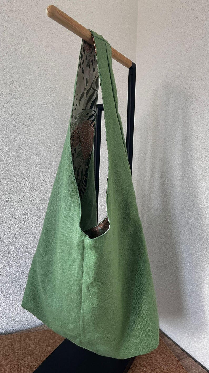May include: A solid sage green hobo bag with a single wide shoulder strap. The interior lining features a contrasting pattern of dark brown and cream botanical leaf illustrations, visible along the inner edge and the top opening of the bag.