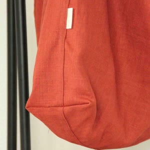 May include: A rust-colored fabric bag with a visible woven texture and a small white rectangular brand label sewn into the side seam. The bag features a structured bottom panel and is suspended from two thin black vertical supports.