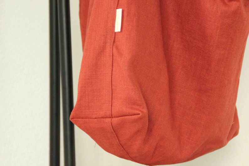 May include: A rust-colored fabric bag with a visible woven texture and a small white rectangular brand label sewn into the side seam. The bag features a structured bottom panel and is suspended from two thin black vertical supports.