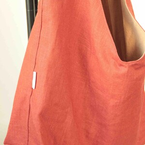 May include: A terracotta-colored hobo bag made of a textured fabric with a visible weave. The bag features a wide shoulder strap, a deep main compartment, and a small white rectangular brand tag sewn into the side seam of the exterior panel.