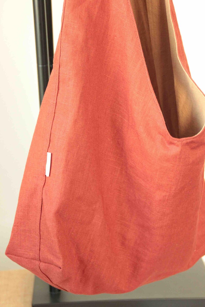 May include: A terracotta-colored hobo bag made of a textured fabric with a visible weave. The bag features a wide shoulder strap, a deep main compartment, and a small white rectangular brand tag sewn into the side seam of the exterior panel.