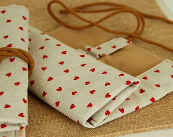 May include: A beige fabric roll-up organizer featuring a repeating pattern of small red hearts. The organizer includes a brown cord for securing the roll and has a smooth, light-colored interior lining. The exterior fabric has a visible woven texture.