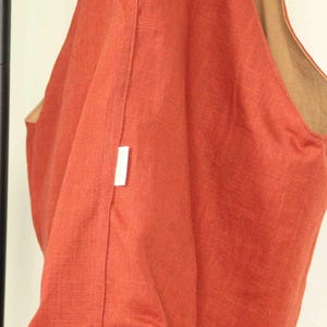 May include: A hobo-style shoulder bag crafted from a textured, rust-orange fabric. The bag features a wide, integrated shoulder strap and a contrasting tan-colored interior lining. A small, rectangular white fabric tag is sewn into the side seam.
