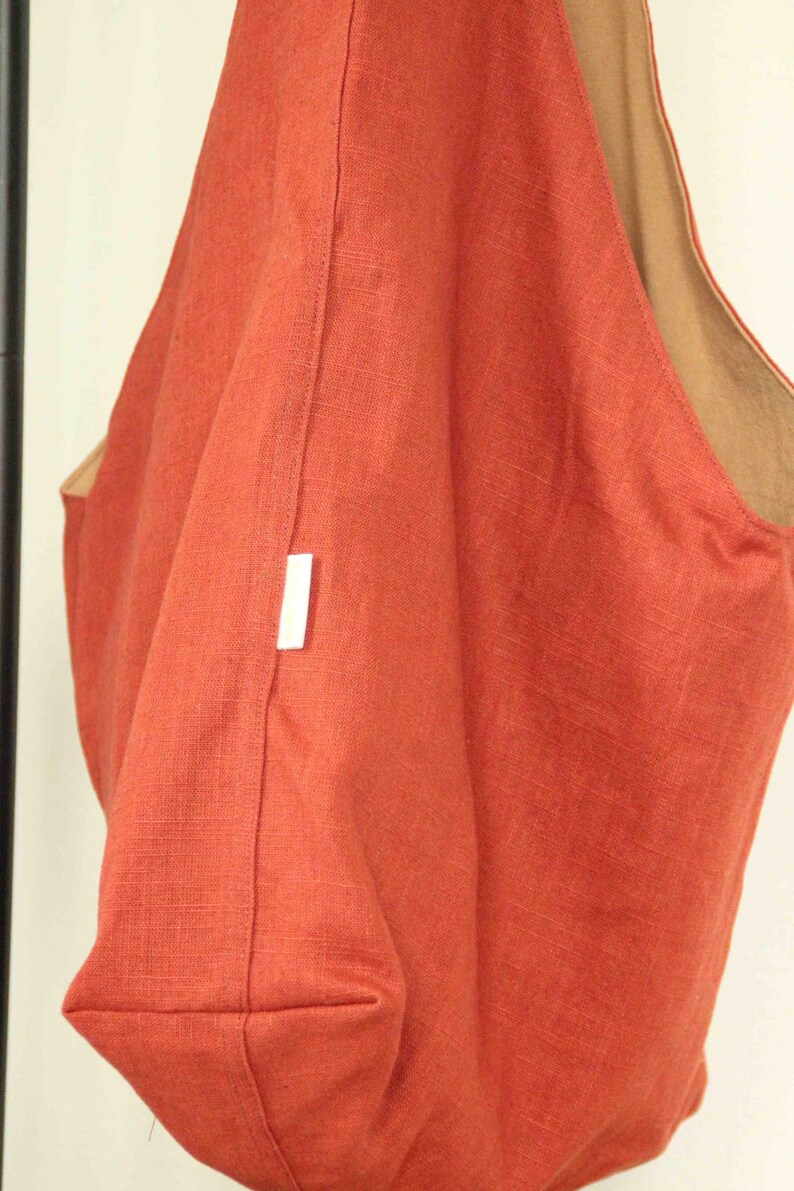 May include: A hobo-style shoulder bag crafted from a textured, rust-orange fabric. The bag features a wide, integrated shoulder strap and a contrasting tan-colored interior lining. A small, rectangular white fabric tag is sewn into the side seam.