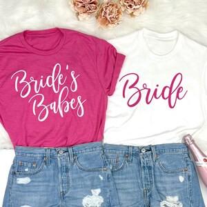 May include: Two white t-shirts with pink and white text. The pink shirt says "Bride's Babes" and the white shirt says "Bride". The shirts are laid out on a white fluffy surface with a pair of denim shorts and a bottle of pink champagne.