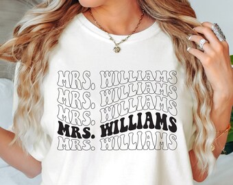 May include: Cream-colored t-shirt with the text "MRS. WILLIAMS" in a retro font. The text is stacked in a wavy pattern, with one instance in black. A casual, everyday wear tee.