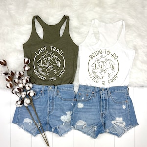 May include: Two tank tops featuring a mountain scene and text. The green tank top reads "Last Trail Before The Veil" and the white tank top reads "Bride-To-Be Wild & Free".