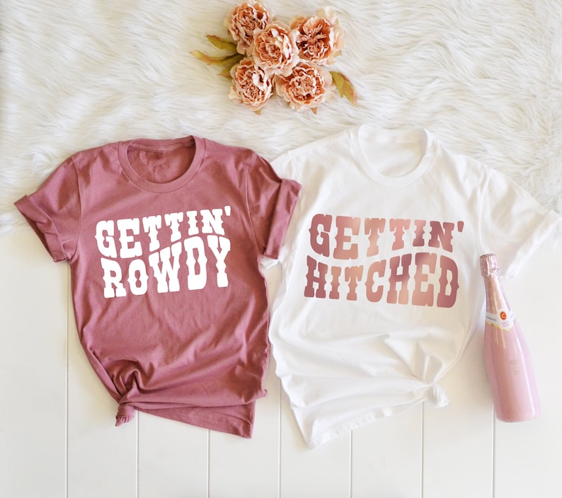 May include: Two white t-shirts with a pink and white ombre design. The text on the shirts reads "Gettin' Rowdy" and "Gettin' Hitched".