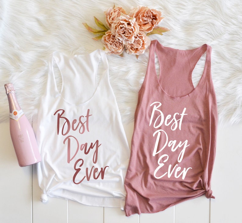 May include: Two white tank tops with the text "Best Day Ever" in rose gold lettering. The tank top on the left is white and the tank top on the right is a light pink color.