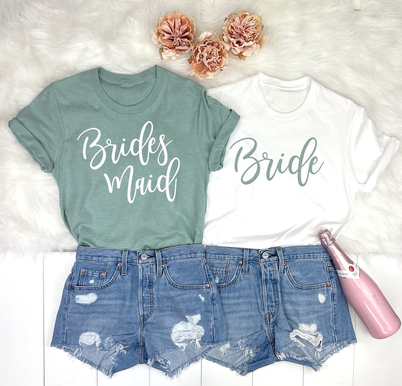 May include: Two white t-shirts with the words "Bride" and "Bridesmaid" written in a light green script font. The shirts are worn over denim shorts and are displayed on a white fuzzy surface.
