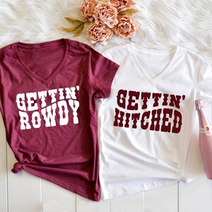 May include: Two white v-neck t-shirts with maroon text. One shirt says "Gettin' Rowdy" and the other says "Gettin' Hitched".