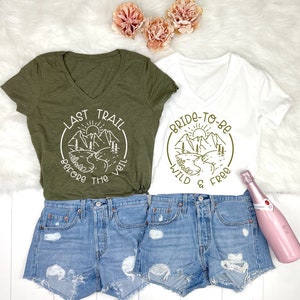 May include: Two green and white t-shirts with a mountain scene and text. The green shirt says "Last Trail Before The Veil" and the white shirt says "Bride-To-Be Wild & Free".