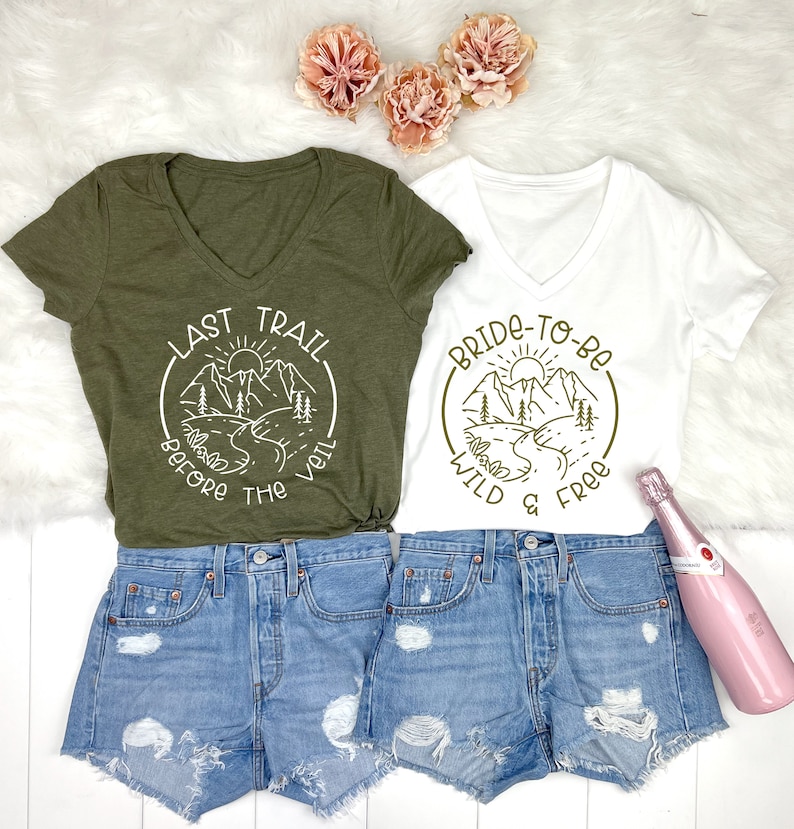 May include: Two green and white t-shirts with a mountain scene and text. The green shirt says "Last Trail Before The Veil" and the white shirt says "Bride-To-Be Wild & Free".
