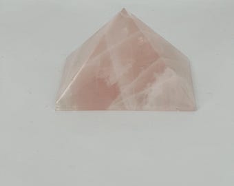 Op de afbeelding: Een piramidevormig rozenkwartskristal. Het kristal heeft een zachtroze kleur met natuurlijke adering en een gepolijste afwerking. De piramide heeft een vierkante basis en loopt taps toe naar een punt. Het is een decoratief item.