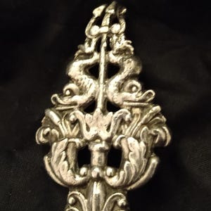 May include: A silver spoon with an ornate, detailed design featuring two figures and floral motifs. The handle is long and slender.