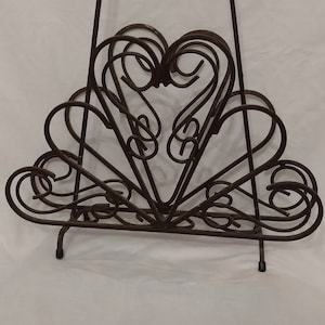 May include: A decorative metal stand with a heart-shaped design. The stand is made of dark brown metal and features a series of swirling, heart-shaped elements. It has a vertical back and two feet.