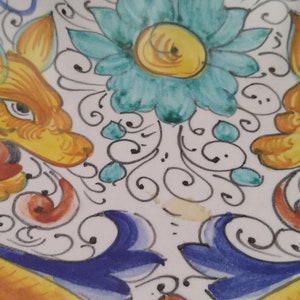 May include: A decorative ceramic plate with a symmetrical design. Two stylized, yellow-orange creatures with blue accents face each other. A turquoise flower and swirling patterns fill the centre, all set against a white background.