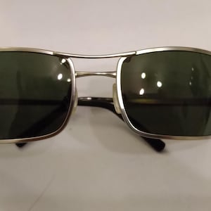 May include: Silver-framed sunglasses with dark green lenses. The sunglasses have a rectangular shape and a slightly curved design.