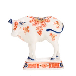 Può includere: Una statuetta di mucca in ceramica bianca su una base rettangolare. La mucca presenta motivi floreali arancioni e dorati, corna blu e una coda blu. La base è decorata con un motivo a griglia arancione, stelle blu e motivi floreali.