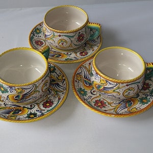 May include: Three ceramic teacups and saucers with a white background and colorful floral and bird designs. The cups have a yellow rim and the saucers have a yellow border.