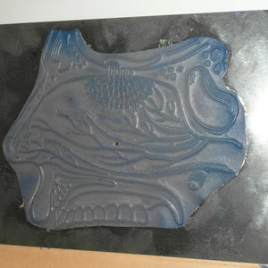 Puede incluir: Un sello de goma azul con un dise&ntilde;o floral ornamentado, montado sobre una placa de metal oscuro. El sello tiene un patr&oacute;n detallado en relieve con varias formas florales y org&aacute;nicas. La placa de metal tiene forma rectangular.