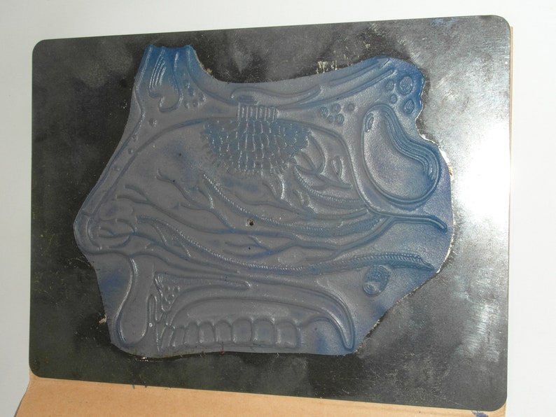 Puede incluir: Un sello de goma azul con un dise&ntilde;o floral ornamentado, montado sobre una placa de metal oscuro. El sello tiene un patr&oacute;n detallado en relieve con varias formas florales y org&aacute;nicas. La placa de metal tiene forma rectangular.