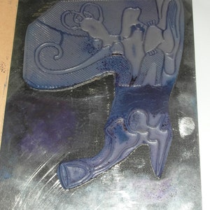 Puede incluir: Un sello de goma azul con un dise&ntilde;o decorativo que presenta una bota y una figura estilizada. El sello est&aacute; montado sobre una base rectangular con un fondo oscuro y un borde claro.