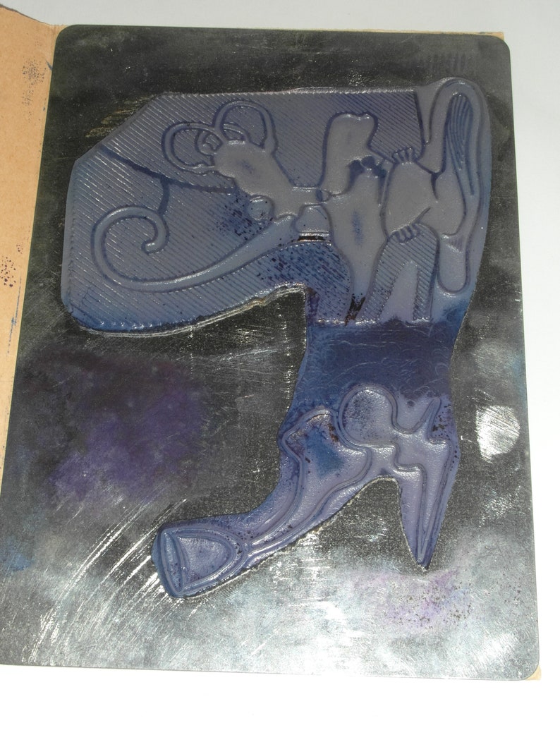Puede incluir: Un sello de goma azul con un dise&ntilde;o decorativo que presenta una bota y una figura estilizada. El sello est&aacute; montado sobre una base rectangular con un fondo oscuro y un borde claro.