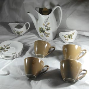 Könnte beinhalten: Ein Vintage-Tee-Set mit einer weißen Teekanne, einer Milchkännchen und einer Zuckerdose mit Blumenmuster. Das Set enthält vier braune Teetassen und Untertassen.