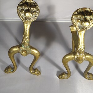 Peut inclure: Paire de chenets anciens en laiton. Chaque chenet présente une tête de lion au sommet, une tige décorative et trois pieds incurvés. Le laiton a une patine chaude et vieillie, avec des zones d'oxydation verte.