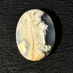 Puede incluir: Gema ovalada con una apariencia nublada y en capas. La piedra presenta una mezcla de tonos blancos, beige y azules, con una zona oscura, casi negra, en un lado. El efecto general recuerda a un paisaje o formación de nubes.