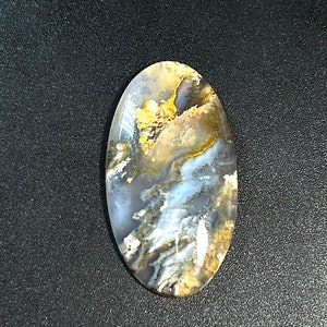 Könnte beinhalten: Ein ovaler, polierter Edelstein-Cabochon mit einer durchscheinenden Basis und wirbelnden Mustern aus grauen, weißen und goldbraunen Einschlüssen. Die Oberfläche ist glatt und reflektierend und zeigt ein komplexes, organisches und marmoriertes Aussehen.