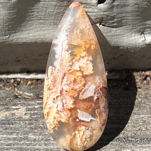 May include: A teardrop-shaped gemstone pendant with a translucent appearance. The stone features a mix of colors, including beige, orange, and white, with a pattern resembling a landscape. The pendant is set against a weathered wooden surface.