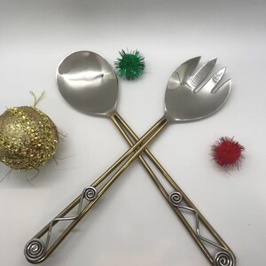 May include: A set of two silver salad serving utensils with gold handles. The handles are decorated with a swirl design. The utensils are crossed over each other.
